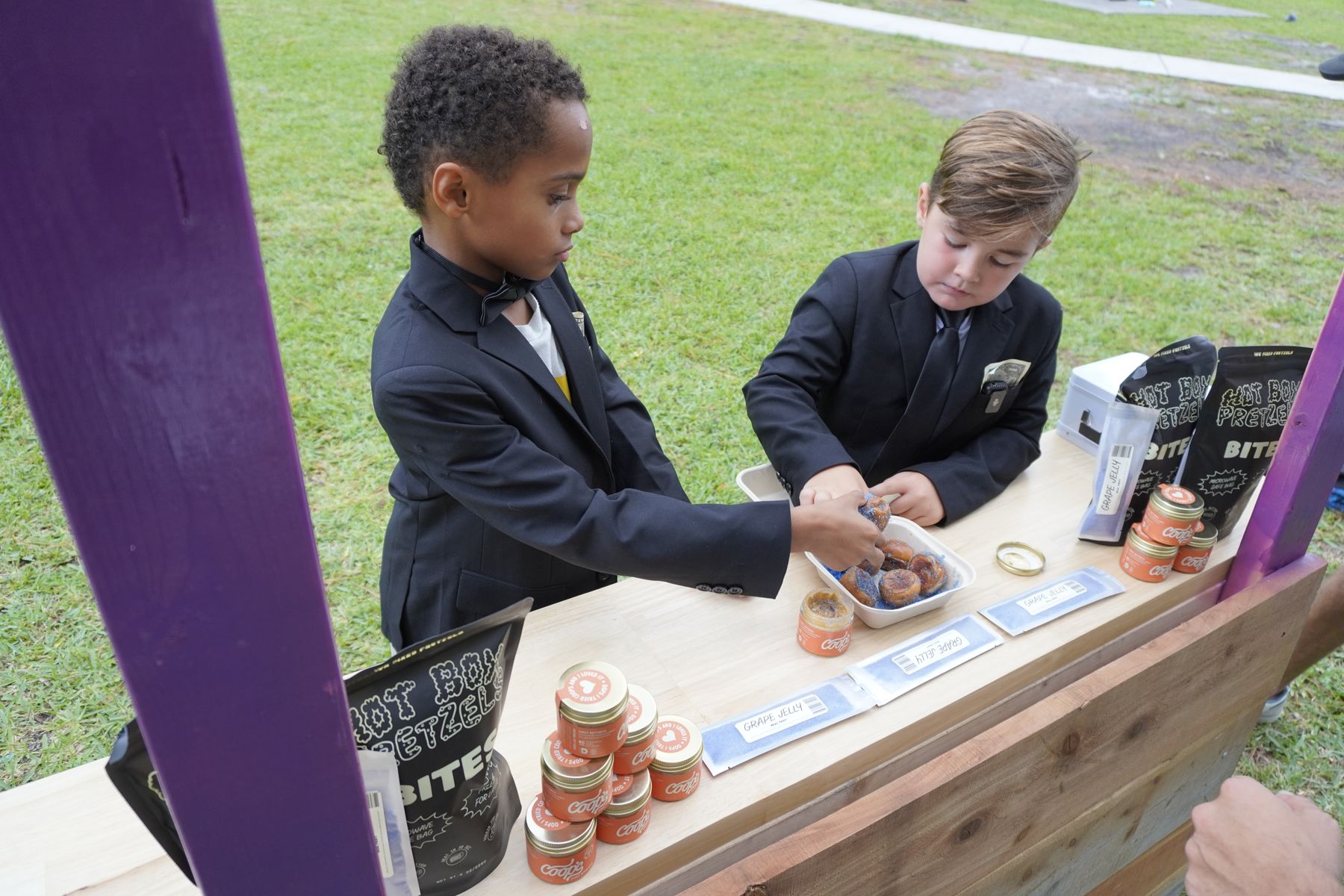 Two kids in suits selling PB&J pretzels at a wooden stand